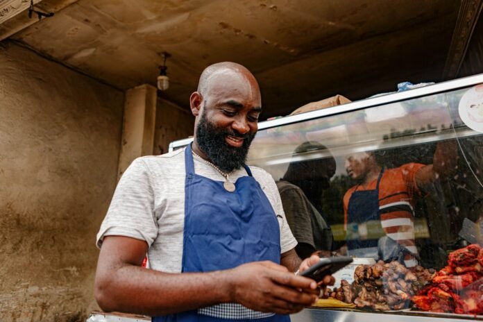 Small business owner in an apron checking his phone, representing entrepreneurship and business registration in Nigeria